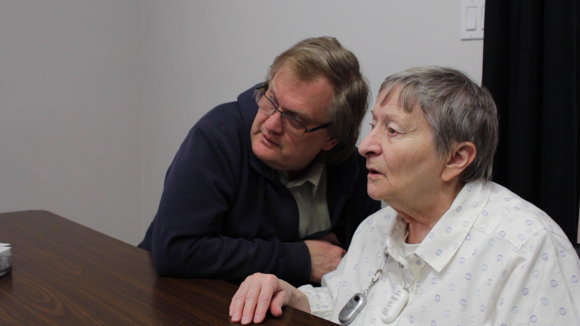 Christine, a white woman in her 70s, wearing a light patterned shir, sitting at a table. Her intervenor, a white man wearing a black hoodie and glasses, leans in close to her.