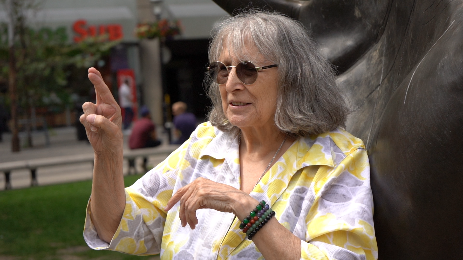 Shari, a white woman in her 70s wearing a yellow and grey blouse, dark glasses, and beaded bracelets. She stands in a park, signing, A statue of a hand is behind her.