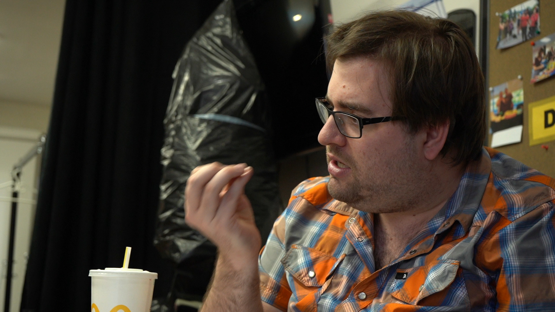 Michael, a white man in his early 30s wearing a plaid orange and blue shirt and black glasses, signs while sitting at a table. A disposable drink cup sits in front of him, and a cork board with photos pinned to itis in the background